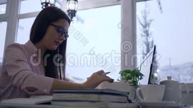 在网上学习，学生女孩使用笔记本电脑，并在笔记本上写笔记，坐在咖啡馆的桌子上，背景是大型的。